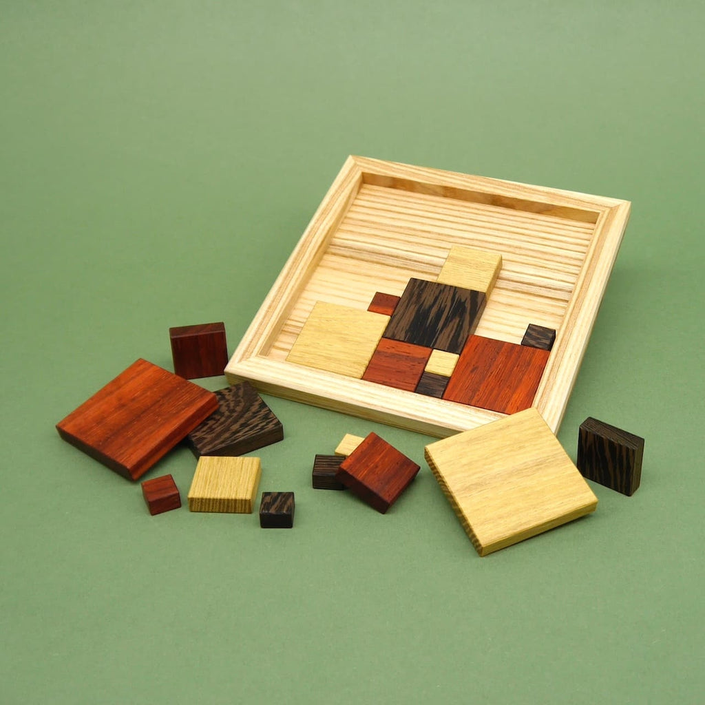 Ein teilweise gelöstes Holzpuzzle mit bunten Holzquadraten in einem Rahmen. Viele Teile liegen verstreut auf dem grünen Untergrund rund um das Spielfeld.