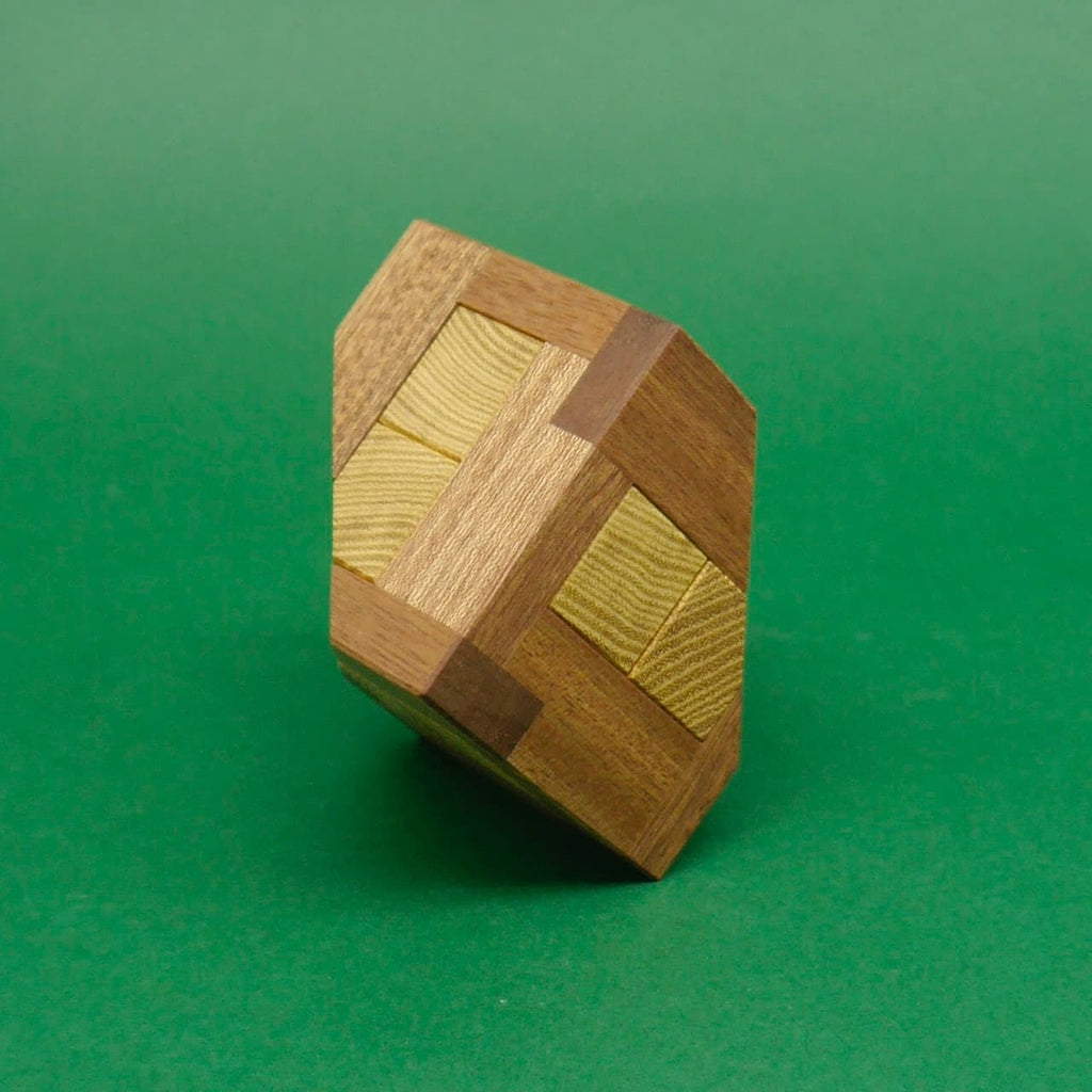 Ein geometrisch präzise gefertigtes Holzpuzzle in Würfelform mit abgerundeten Kanten, stehend auf einer Spitze vor grünem Hintergrund.