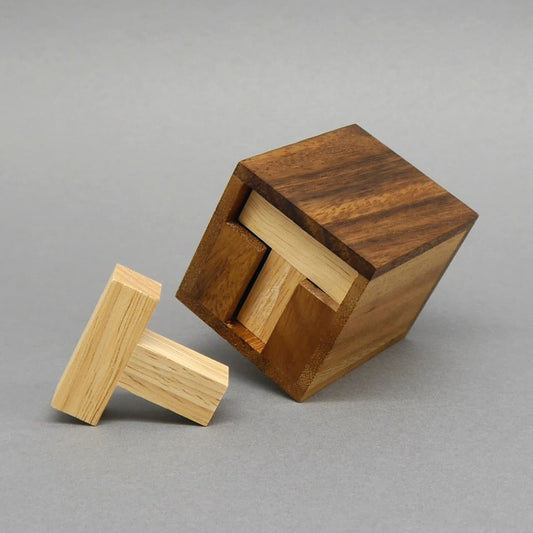 Braune Holzbox kippt auf den grauen Untergrund; zwei helle, T-förmige Holzsteine ragen heraus, einer liegt davor – zeigt das teilweise gelöste „Six T“-Packpuzzle.