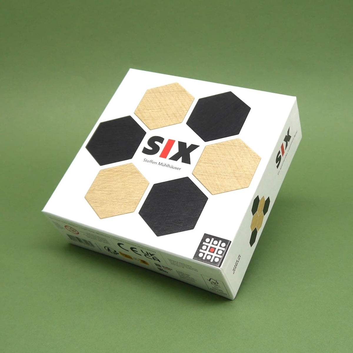 Box des legespiel „SIX“ mit schwarzen und naturfarbenen Sechseck-Holzplättchen auf grünem Hintergrund.