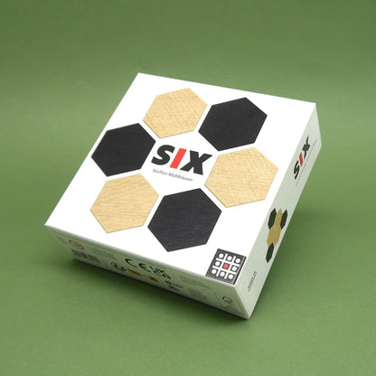 Box des legespiel „SIX“ mit schwarzen und naturfarbenen Sechseck-Holzplättchen auf grünem Hintergrund.
