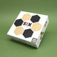 Box des legespiel „SIX“ mit schwarzen und naturfarbenen Sechseck-Holzplättchen auf grünem Hintergrund.