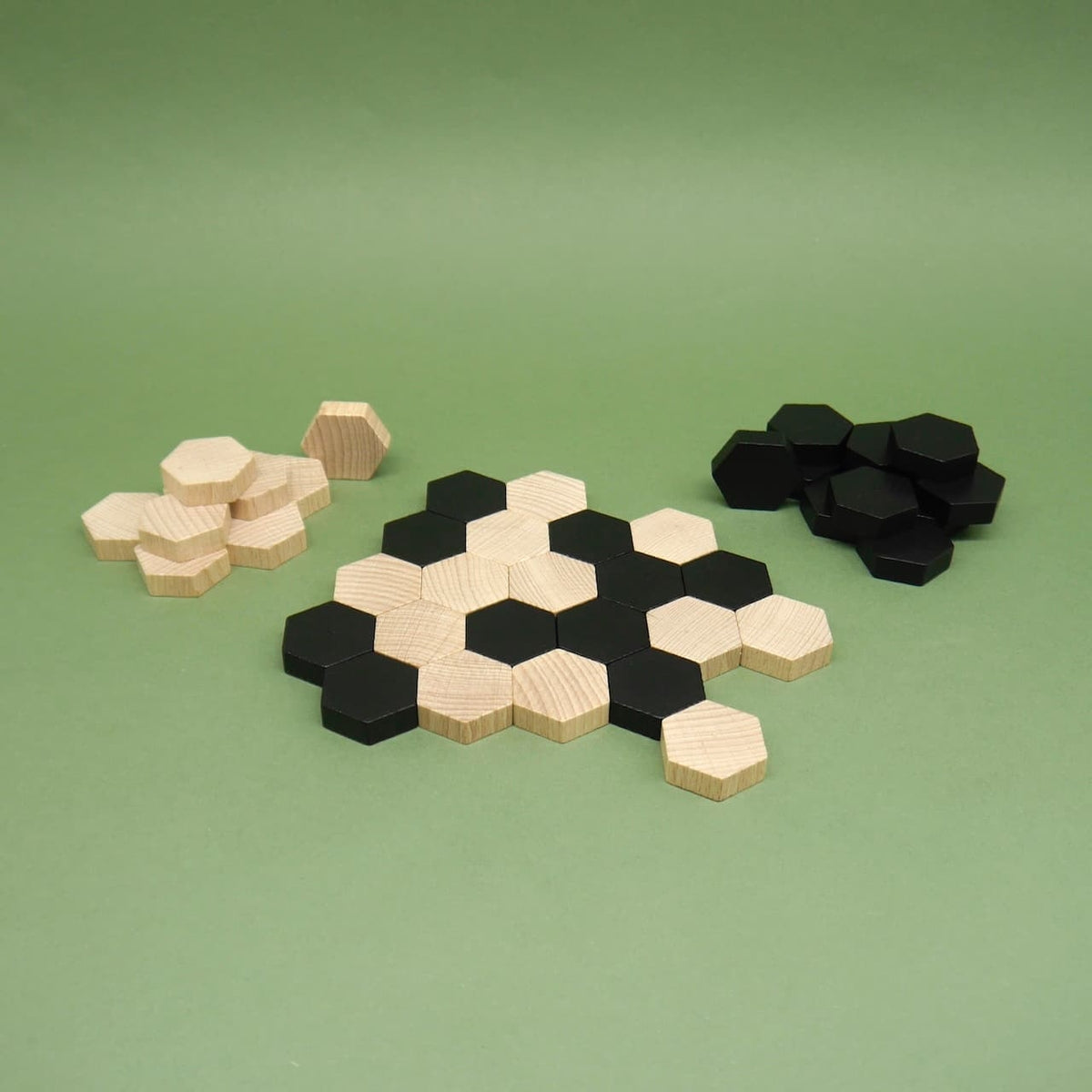 Muster aus Holzhexagonen des gesellschaftsspiel „SIX“, Steine in Natur und Schwarz, Vorratsstapel daneben auf grünem Untergrund.