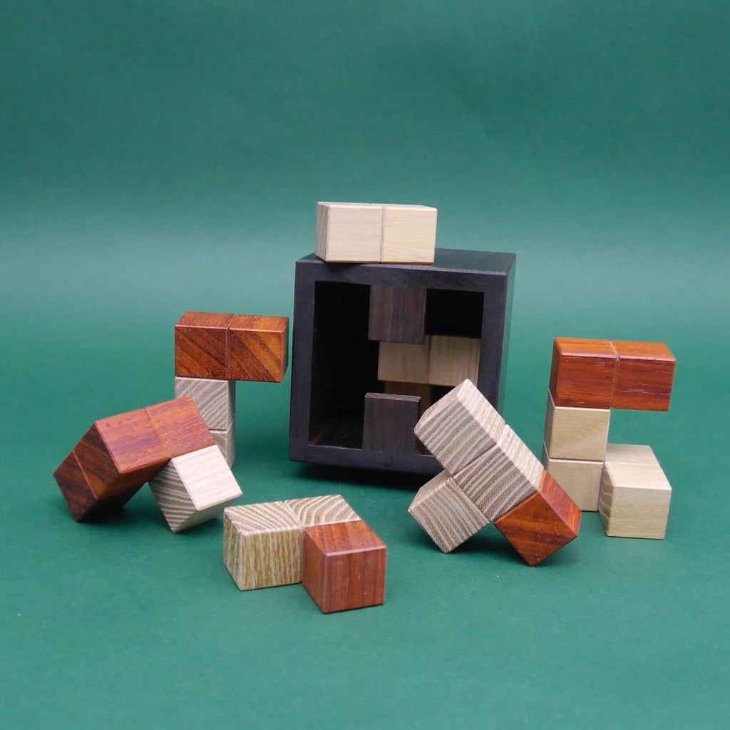 Knobelspiel aus unterschiedlich gefärbten Holzklötzchen vor grüner Fläche, einige Teile stehen, andere liegen vor einer leeren schwarzen Box.