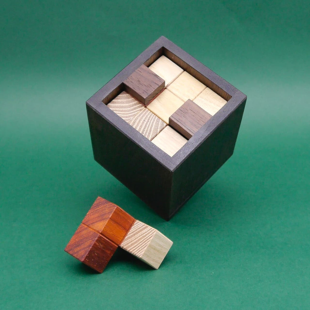 Soma Würfel in dunkler Holzbox vollständig zusammengesetzt, ein weiteres Holzelement liegt davor auf grünem Hintergrund.