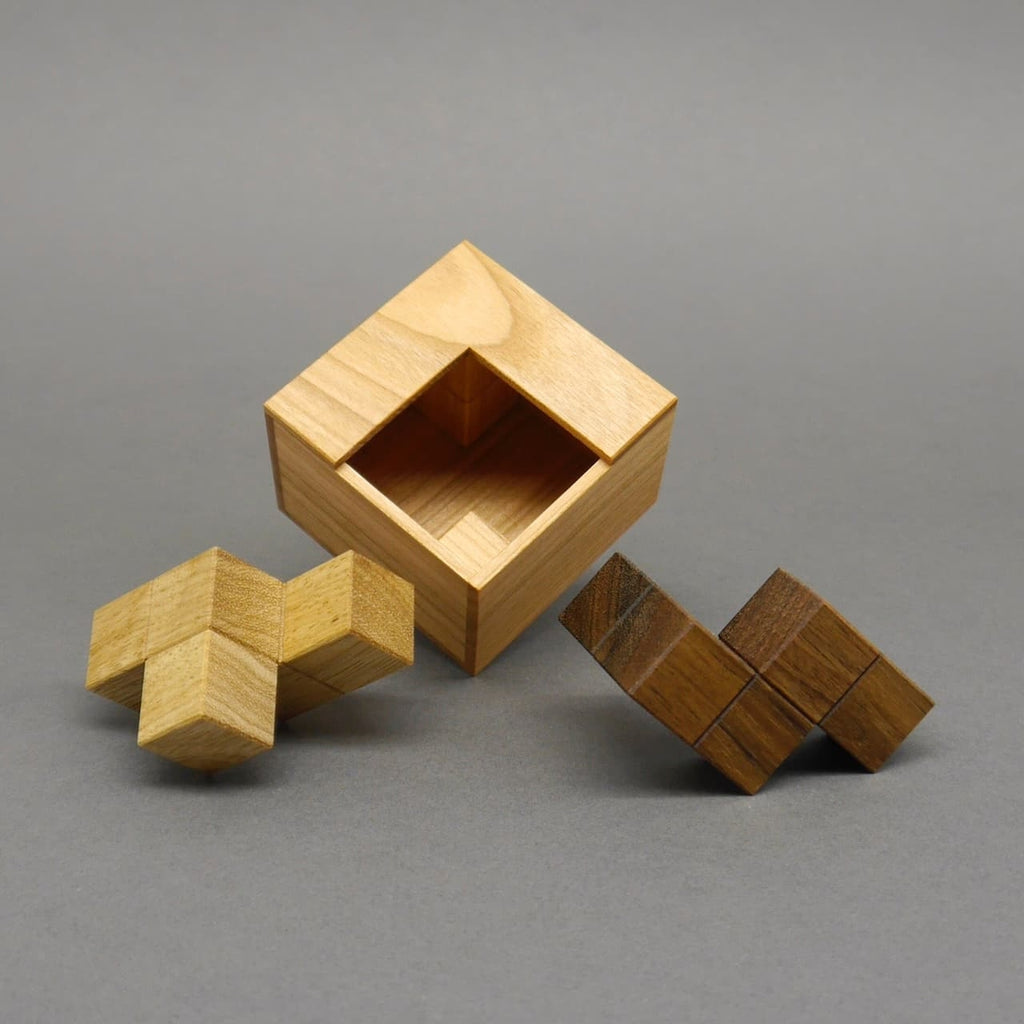 Zwei herausgenommene Holzpuzzle-Teile neben einer leeren Kiste als Teil eines räumlichen Packrätsels.