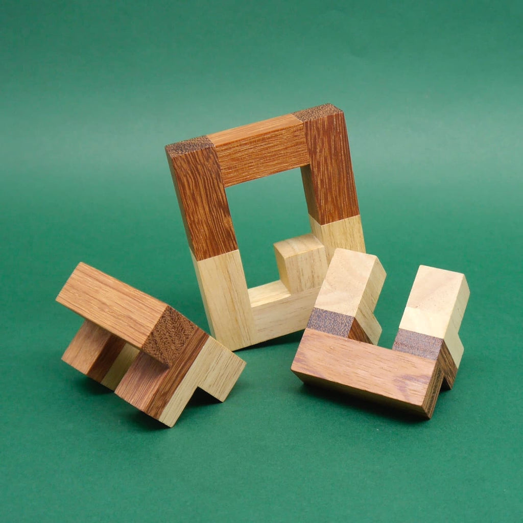 Zerlegtes Logikpuzzle aus hellen und dunklen Holzklötzen; ein rechteckiger Rahmen und zwei Winkelteile liegen auf grünem Untergrund.
