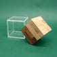 Kippender Holz‑Puzzle‑Würfel ragt halb aus seiner klaren Acrylbox vor grünem Hintergrund heraus