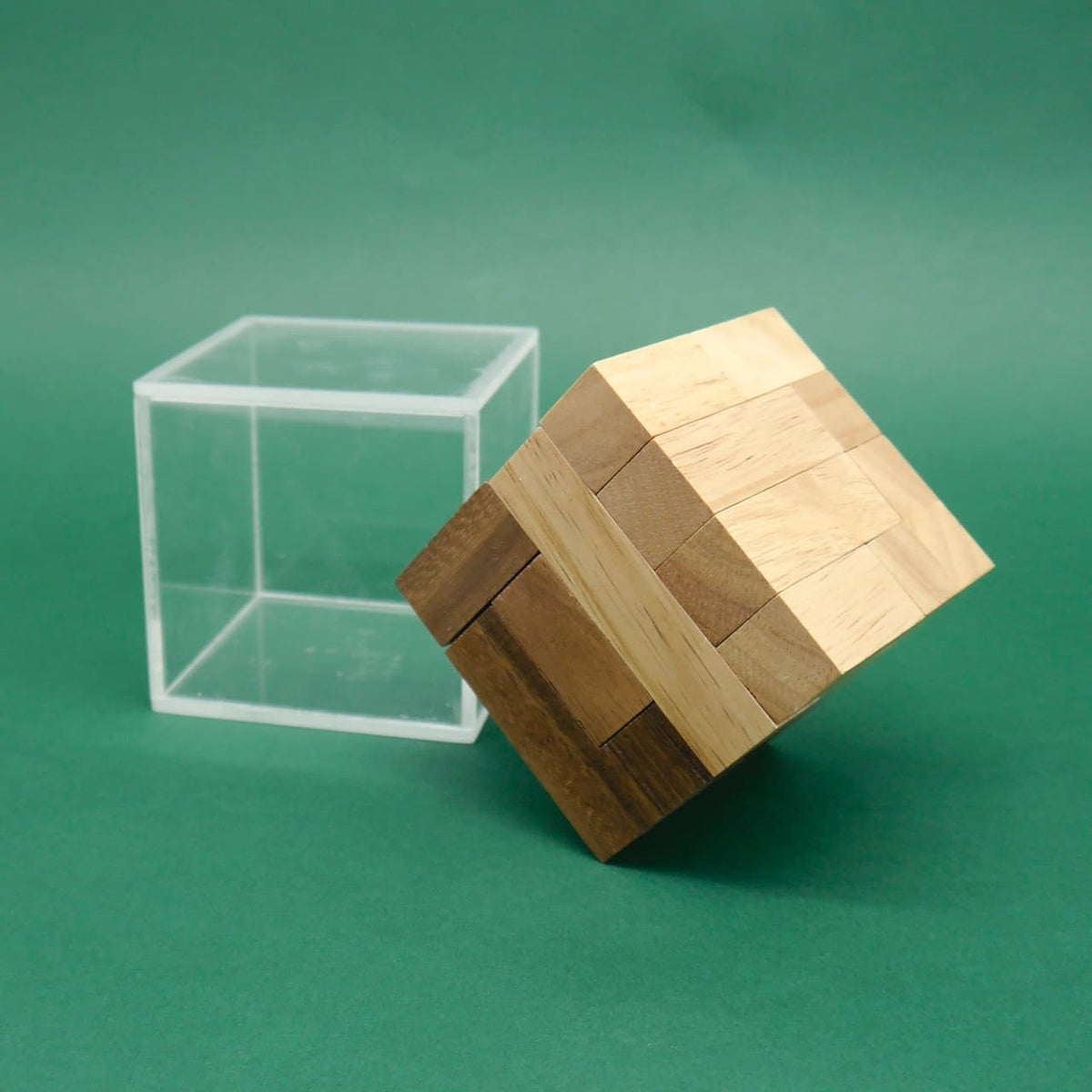 Kippender Holz‑Puzzle‑Würfel ragt halb aus seiner klaren Acrylbox vor grünem Hintergrund heraus
