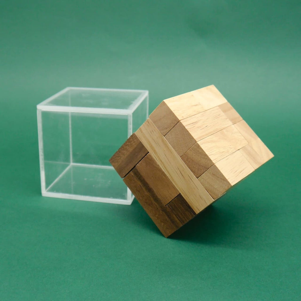 Kippender Holz‑Puzzle‑Würfel ragt halb aus seiner klaren Acrylbox vor grünem Hintergrund heraus