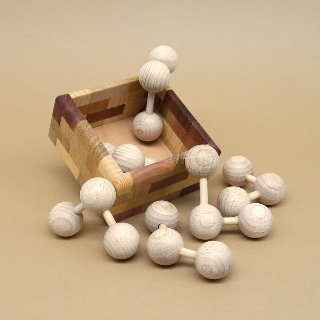 Einzelne Kugel-Stab-Elemente des Holzpuzzle liegen vor gekippter Box; dynamische Anordnung, matte Lichtstimmung auf neutralem Grund.