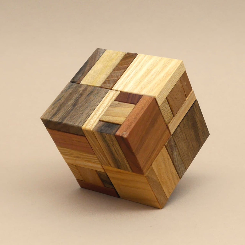 Ein zusammengesetzter Holzwürfel aus verschiedenfarbigen Holzstücken steht leicht gedreht auf einer beige-farbenen Fläche.