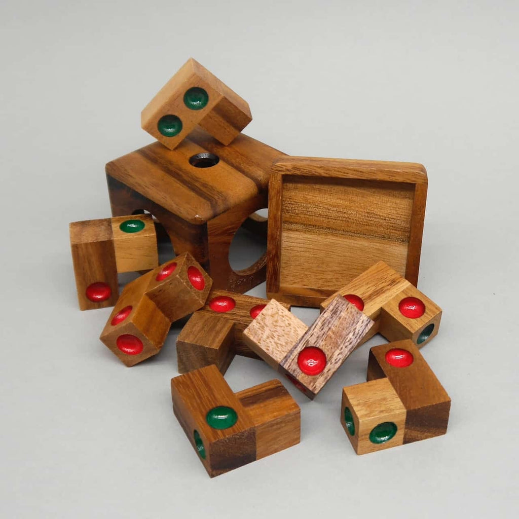 Zerlegte Geduldsspiel-Holzspielteile mit roten und grünen Vertiefungen liegen vor einer quadratischen Box und ihrem Deckel auf grauem Hintergrund.