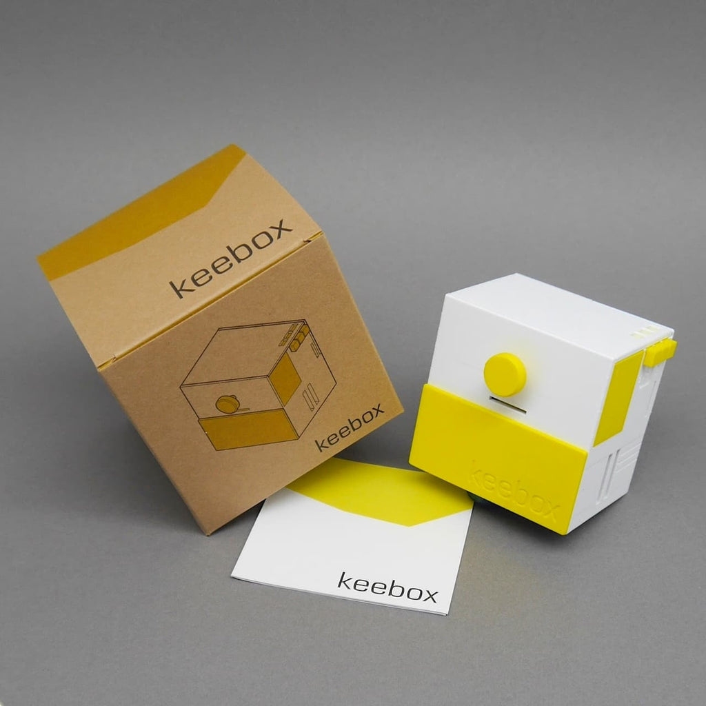Keebox ‚yellow‘ neben geöffneter Schachtel und Faltblatt; Rätselbox in Weiß-Gelb im Dreiviertelprofil.